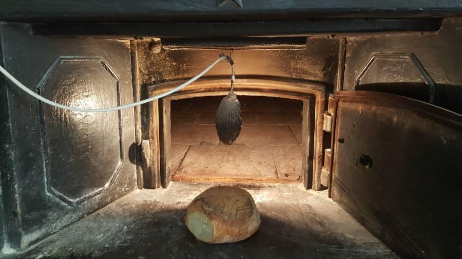 A loaf of bread inside a rustic looking oven with a lightbulb hanging above it. Darkly lit photo with a bright and warm colored light from the bulb shining on the bread.