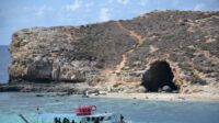 A foreground of very saturated blue sea with a small island in the back and a cave opening into it.