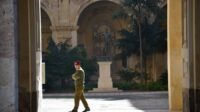 A guard walking on the courtyard of a palace, pillars and vegetation seen in the back