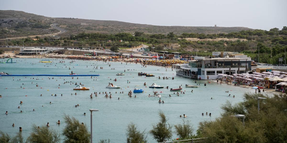 A populated beach in Mellieha in good weather with blue skies, paddleboats and many people in the sea and on land.