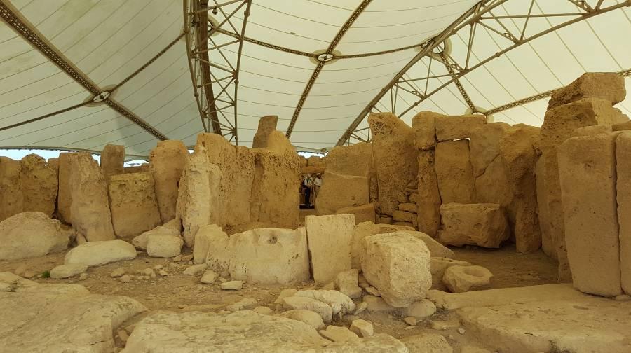 Mnajdra temple complex, temple remains under a protective tent. Tan colored stone in irregular cuts set in  pattern reminiscent of a spacious structure. 