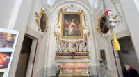 An upwards view of an altar, white walls in the chapel with decadent framing of a painting situated in the centre of the image