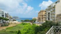 A perspective image of Balluta Bay from a far away viewpoint with greenery and a fraction of the city skyline visible.