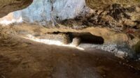 The interior of a cave with a stone wall on the opposite side and the entrance letting in sunlight