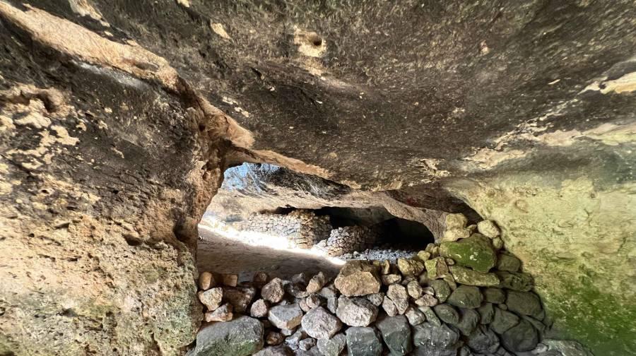 A stone wall with a section made of smaller rounded stones with an opening into a neighboring section of the cave with more constructed stone walls