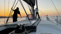 A small white sailboat and two silhouettes standing in front of it, with a sunset at the back