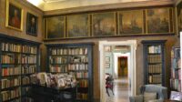 A 16th century green and brown accented library inside a decadent interior of a palace. A white hall into the rest of the palace is seen from a white doorway.