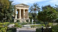 A garden with a pond and Greek reminiscent structure with pillars. Greenery all around, with palms framing the structure