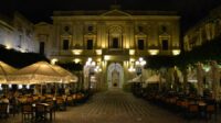 The Maltese National Library with a statue in front during nighttime. Scarcely lit and accompanied by two sets of outside cafe umbrellas on either side.