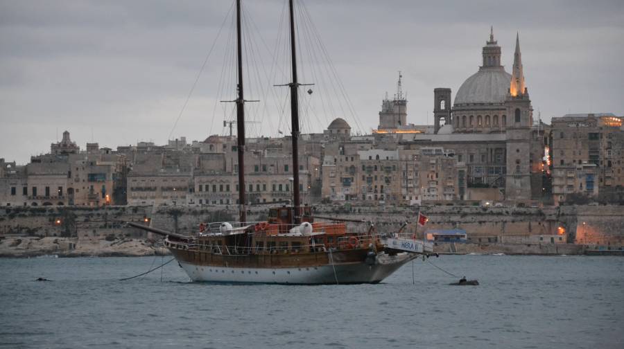 A skyline of Valletta in the evening hours with moody lightinh in the corners of the city. An old style sailing boat in front.