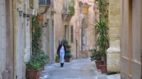 A curving street in Birgu with faint sunlight illuminating it, greenery spilling over the walls and a person walking down the street carrying bags