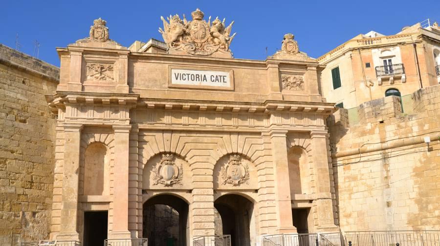 Victoria Gate in Valletta as seen from ground level. An intricate limestone wall with a crest atop an arch-like construction and a sign that reads "Victoria Gate"